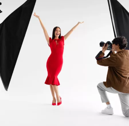Behind the scenes of photoshoot. Elegant female model posing for professional fashion photographer, shooting over white background. Stylish magazine photoshoot