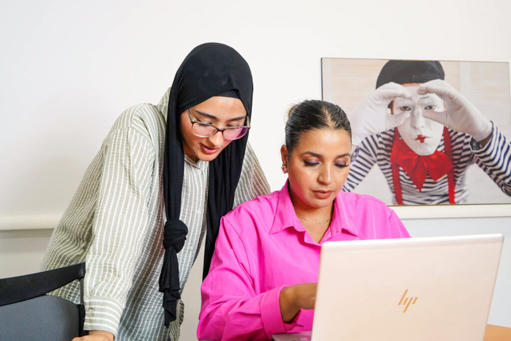 Deux femmes collaborent à un ordinateur portable dans une Agence de Communication Digitale et Création Site web ; l'une est debout, vêtue d'un hijab et de lunettes, l'autre est assise, vêtue d'une chemise rose vif. Une peinture représentant un mime orne le mur derrière elles.