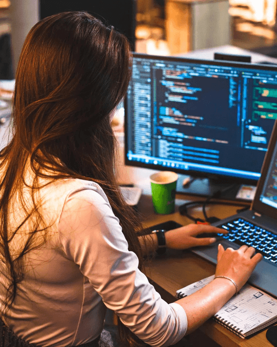 Une personne aux longs cheveux bruns est assise à un bureau, tapant sur un ordinateur portable dont le code est affiché sur deux moniteurs. Un cahier, un stylo et une tasse verte sont posés sur le bureau. L'espace de travail est bien éclairé et organisé.